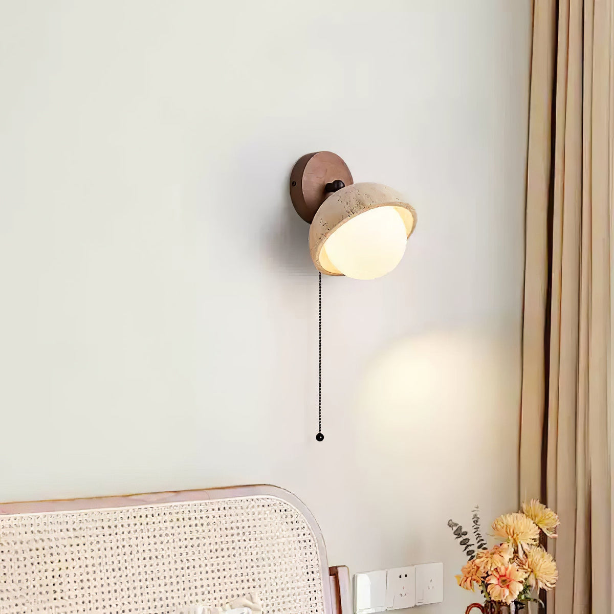 Close crop of a travertine dome wall light from Clérachic, lit above a cane headboard, with a bunch of orange flowers at the edge.