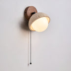 Angled view of the walnut backplate, travertine dome and glowing white globe of this Clérachic wall light, with a black pull chain.