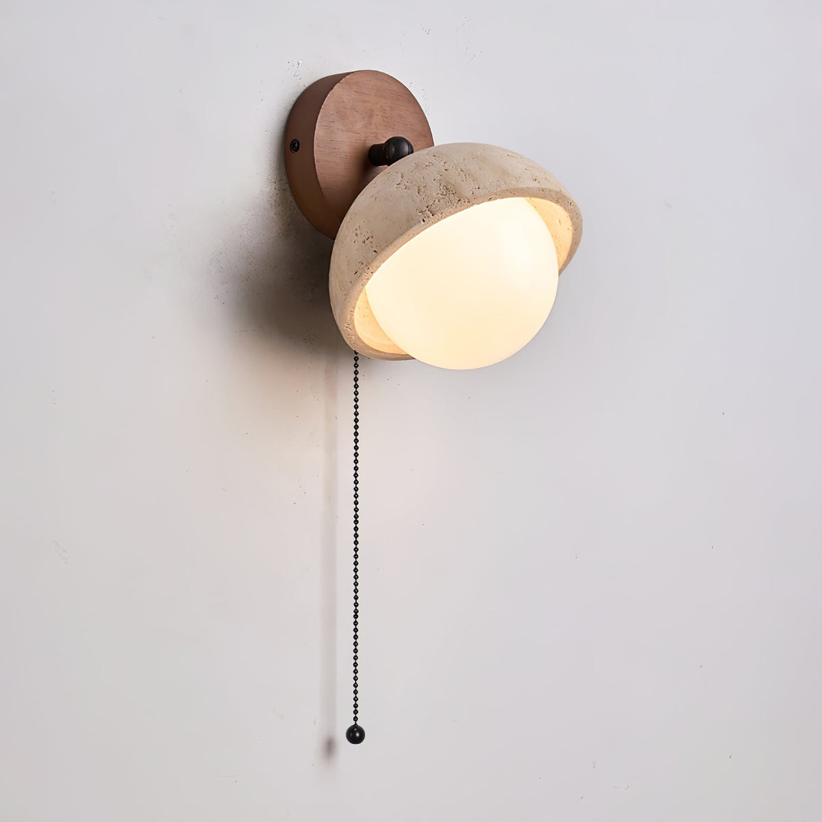 Angled view of the walnut backplate, travertine dome and glowing white globe of this Clérachic wall light, with a black pull chain.
