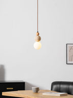 A Clérachic pendant with a wood bead, a travertine ball and a glowing white globe, hung against a pale wall above a wooden table and black chair.