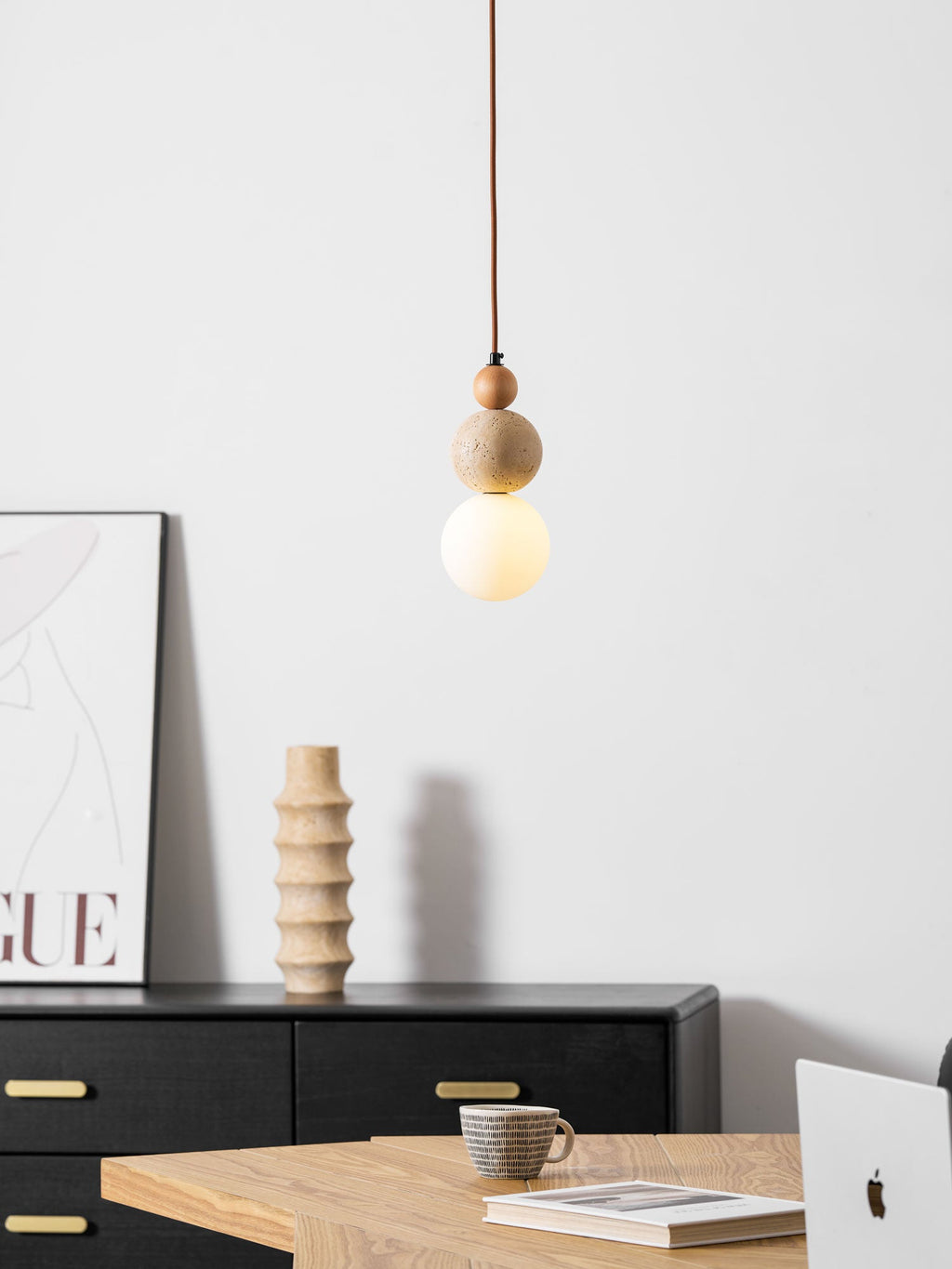 A lit Clérachic pendant with a travertine ball and white globe, in front of a black sideboard, a ribbed vase and a framed poster.