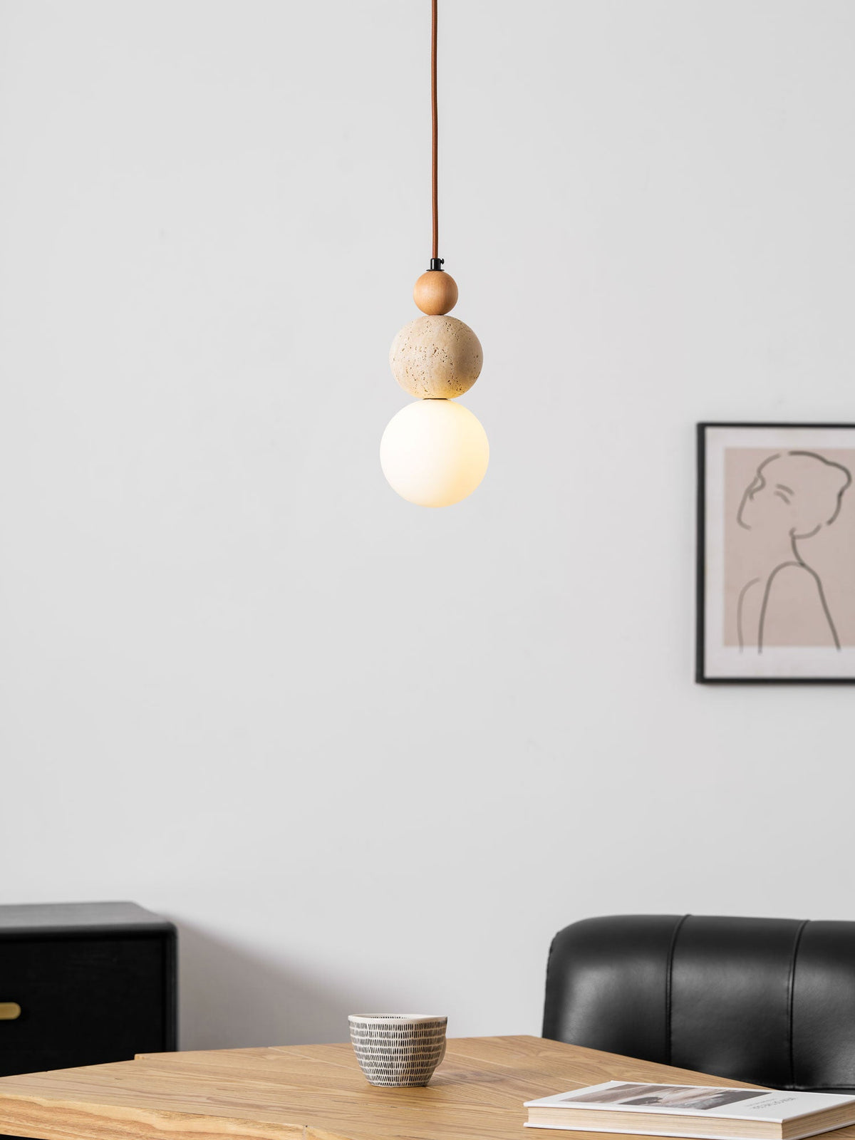 A Clérachic pendant with a wood bead, travertine ball and glowing white globe, beside a framed line drawing of a woman's profile.