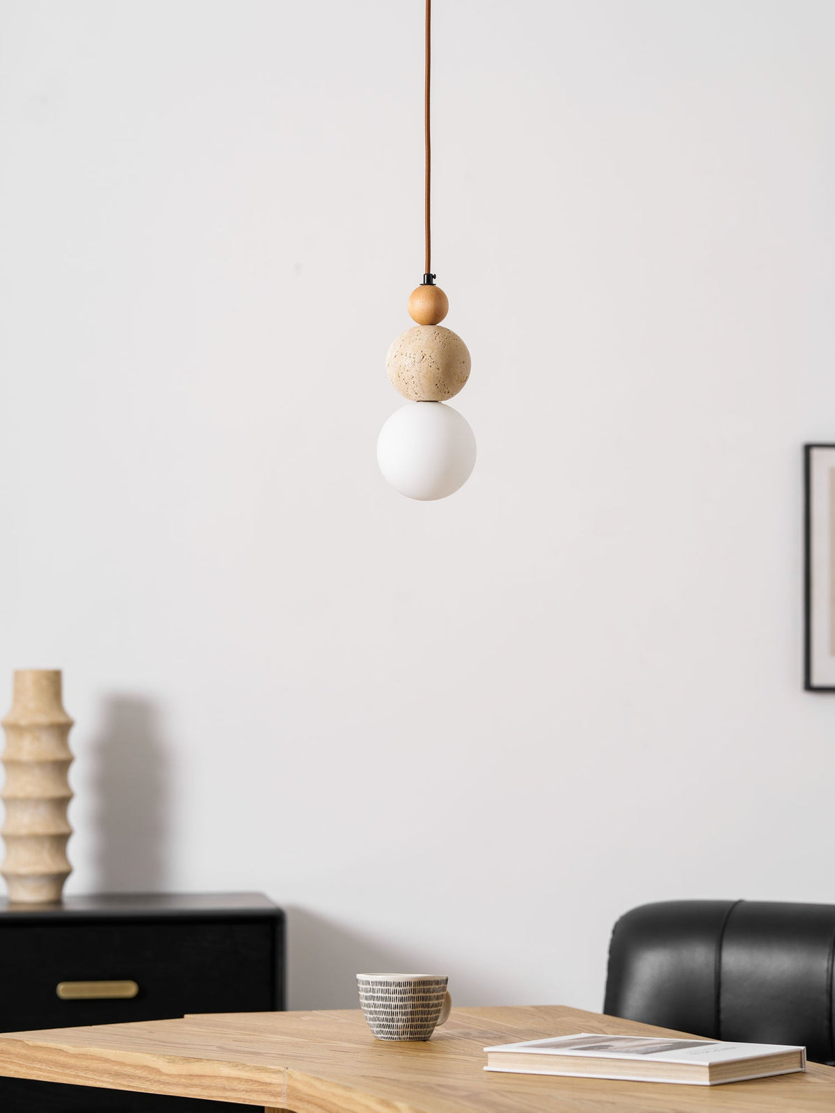 An unlit Clérachic pendant with a travertine ball and white globe, above a wooden table, with a ribbed vase and a black sideboard behind.