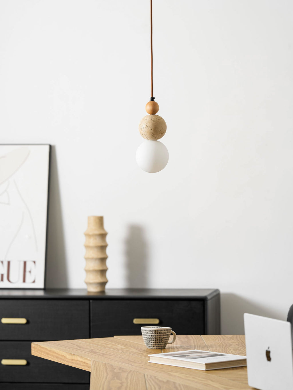 An unlit Clérachic pendant hung in front of a black sideboard with brass handles, a ribbed vase, a poster and a cup on a wooden table.