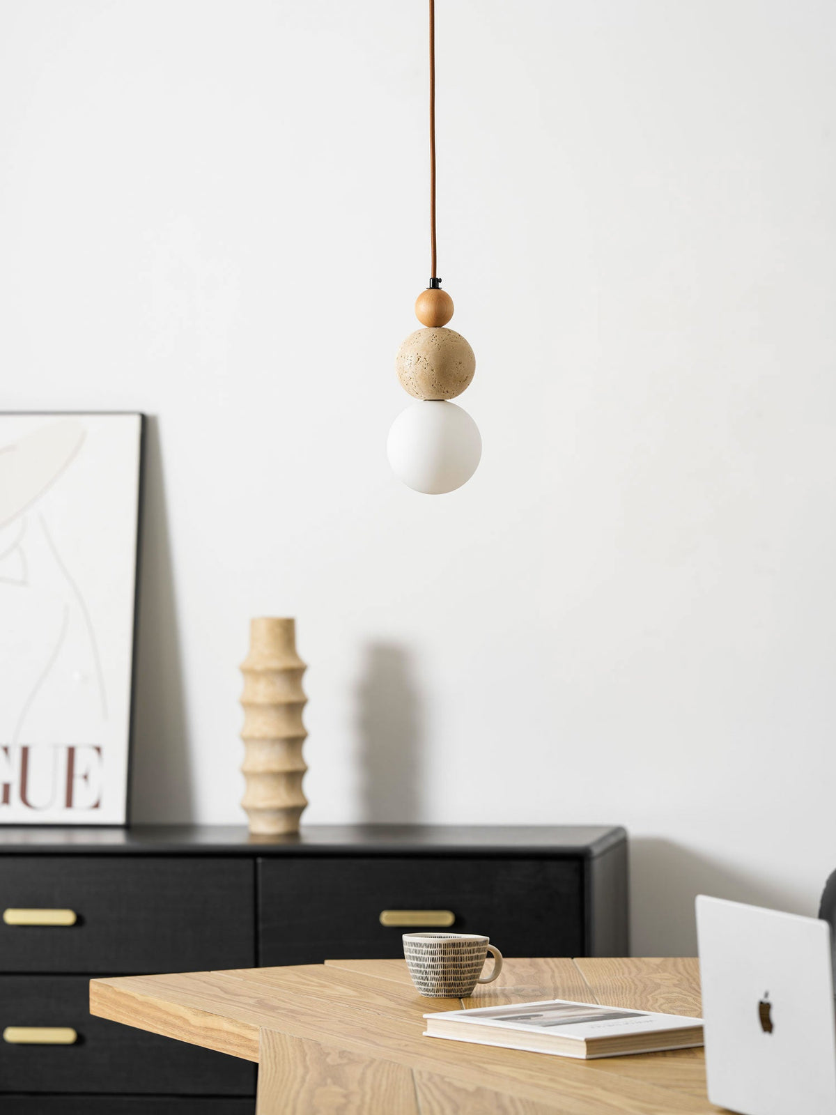 An unlit Clérachic pendant hung in front of a black sideboard with brass handles, a ribbed vase, a poster and a cup on a wooden table.