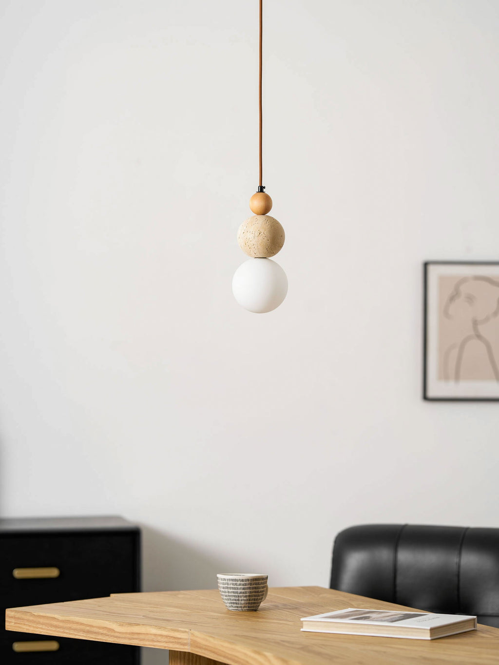A Clérachic pendant with a wood bead, travertine ball and unlit white globe, against a pale wall with a framed line drawing.