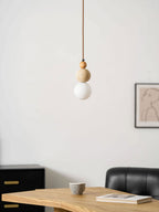 A Clérachic pendant with a wood bead, travertine ball and unlit white globe, against a pale wall with a framed line drawing.