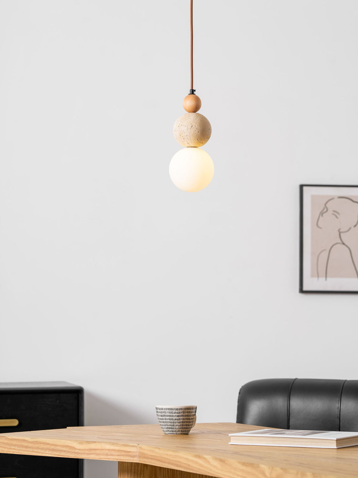 A Clérachic pendant glowing softly, hung beside a framed line drawing, with a patterned cup and a book on a wooden table below.