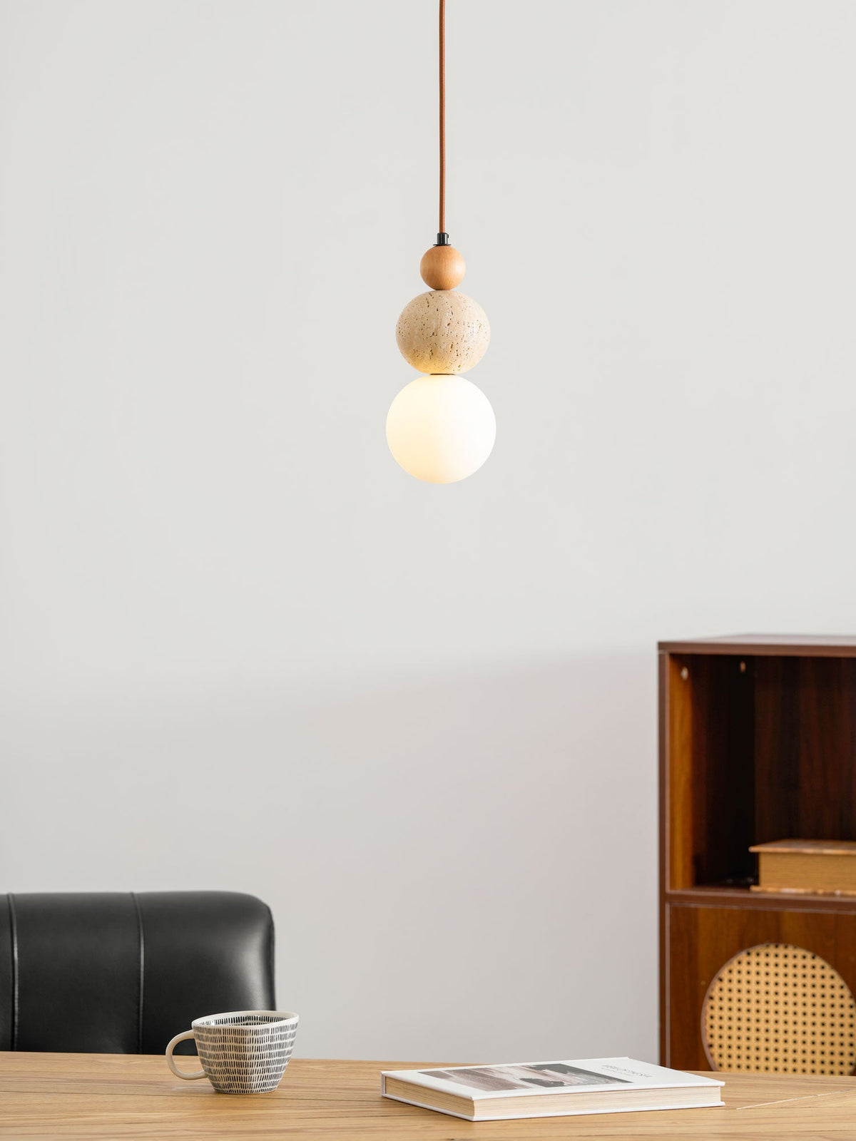 A lit Clérachic pendant with a wood bead, travertine ball and white globe, near a rattan-fronted cabinet and a black leather chair.