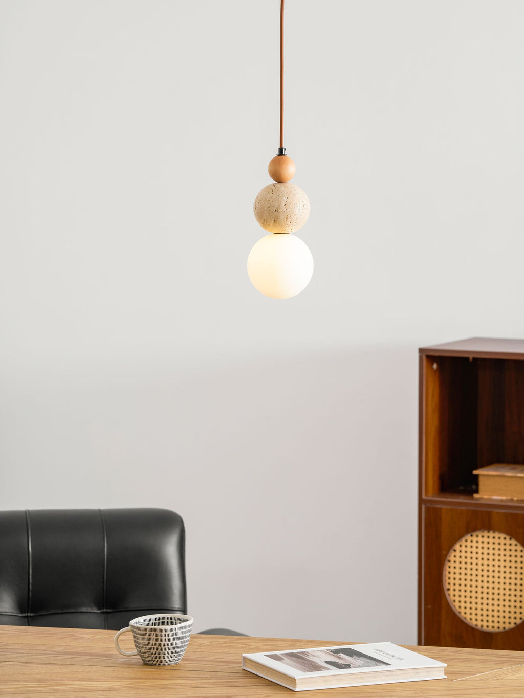 A Clérachic pendant with a travertine ball and a softly lit white globe, above a wooden table with a black chair and a cup of tea.