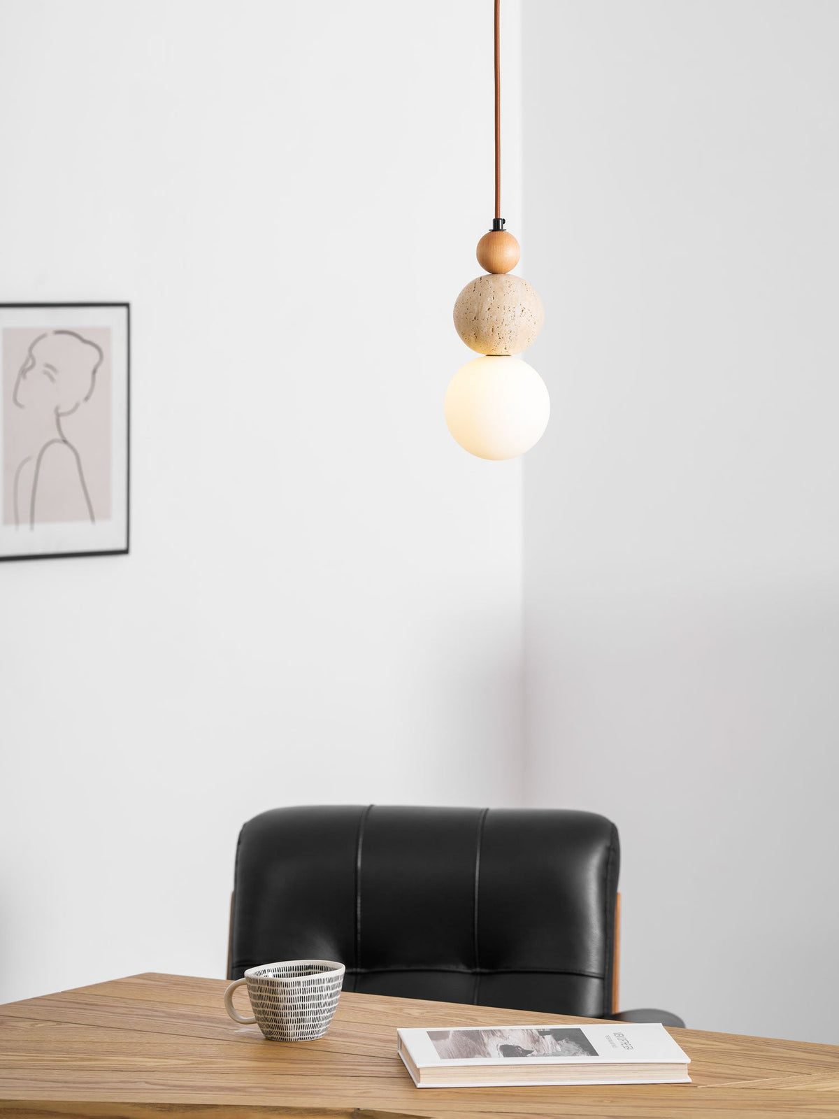 A lit Clérachic pendant beside a framed line drawing, above a wooden table with a black leather chair, a cup and a book.