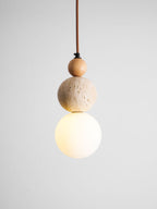 A lit Clérachic pendant with a wood bead, a pitted travertine ball and a glowing white globe, on a pale grey backdrop.