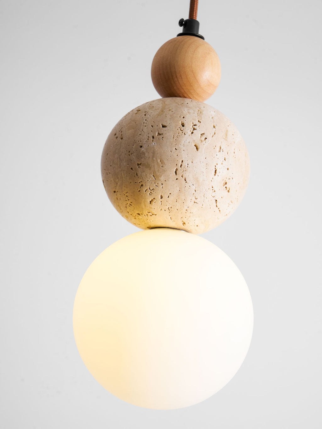 Close view of the lit white globe, the pitted travertine ball and the wood bead on this Clérachic pendant, on a grey backdrop.