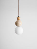 An unlit Clérachic pendant with a small wood bead, a travertine ball and a white globe hanging on a brown cord against grey.