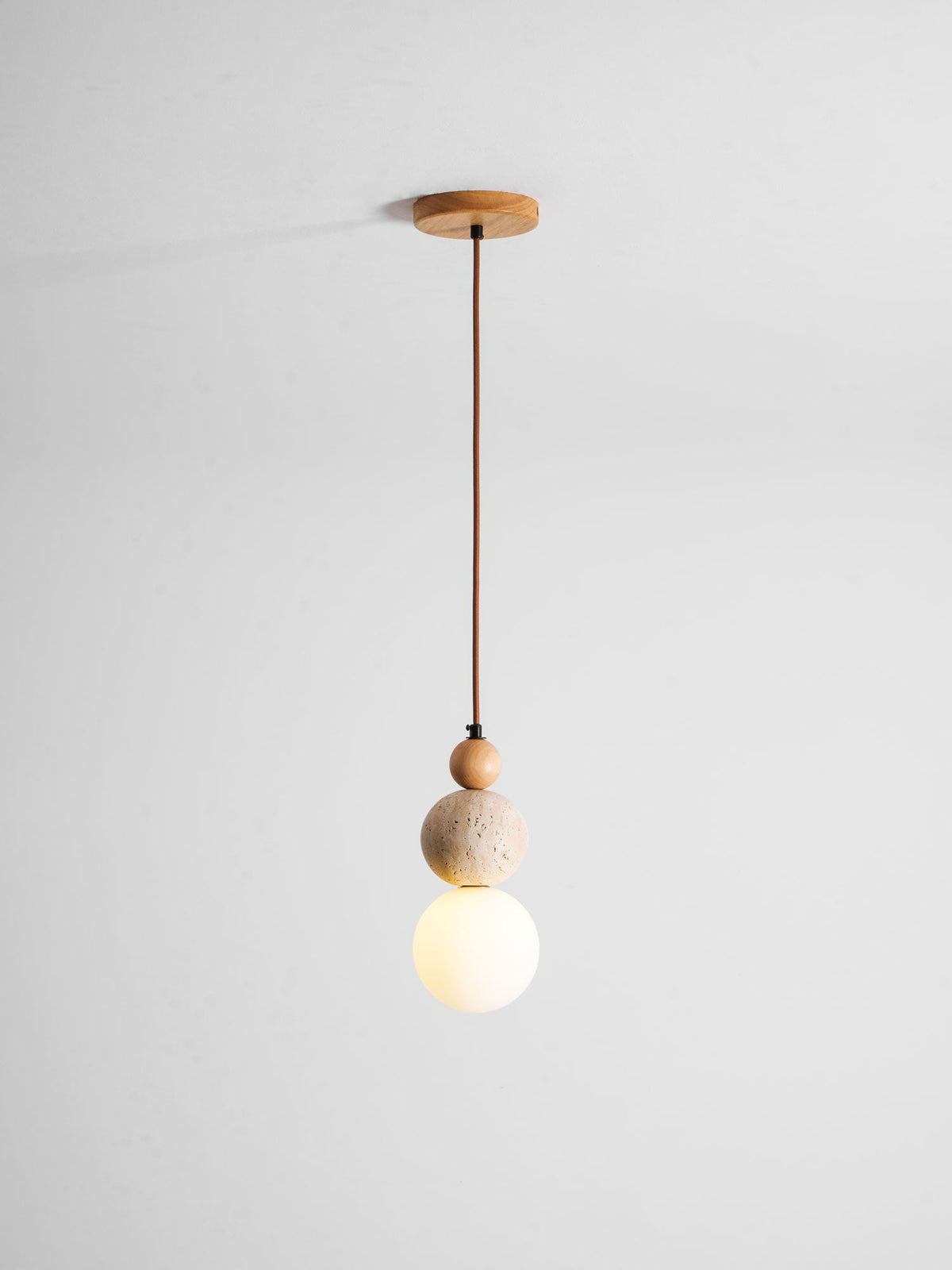 A Clérachic pendant on a brown cord from a round wood canopy, with its white globe glowing warm under the travertine ball.