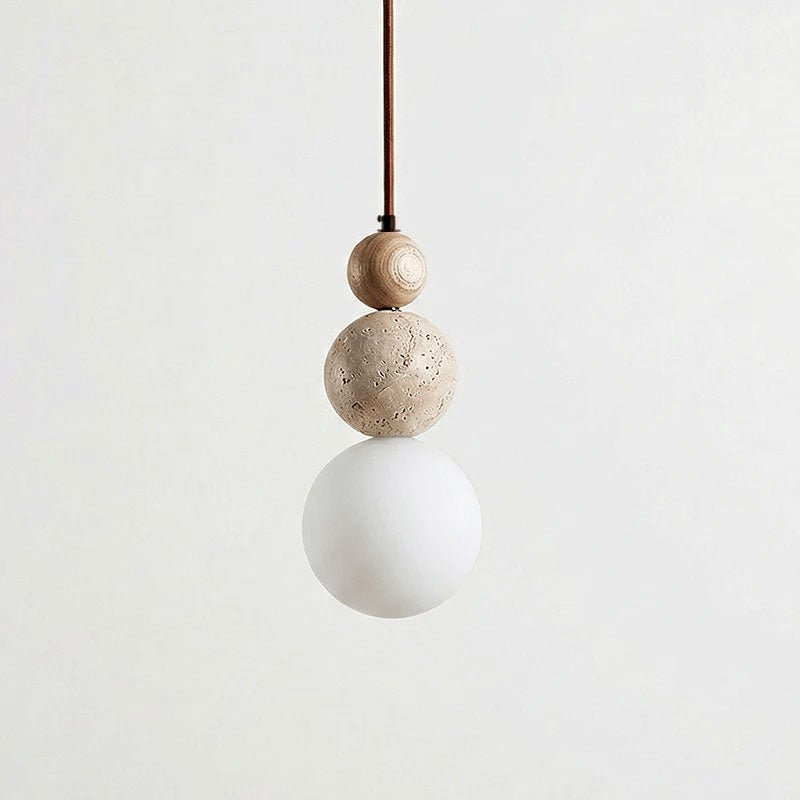A Clérachic pendant hanging on a brown cord, with a wood bead, a travertine ball and a matt white globe against a pale backdrop.