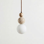 A Clérachic pendant hanging on a brown cord, with a wood bead, a travertine ball and a matt white globe against a pale backdrop.