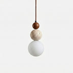 A Clérachic pendant on a plain light backdrop, with a dark walnut bead above a pitted travertine ball and a matt white globe.
