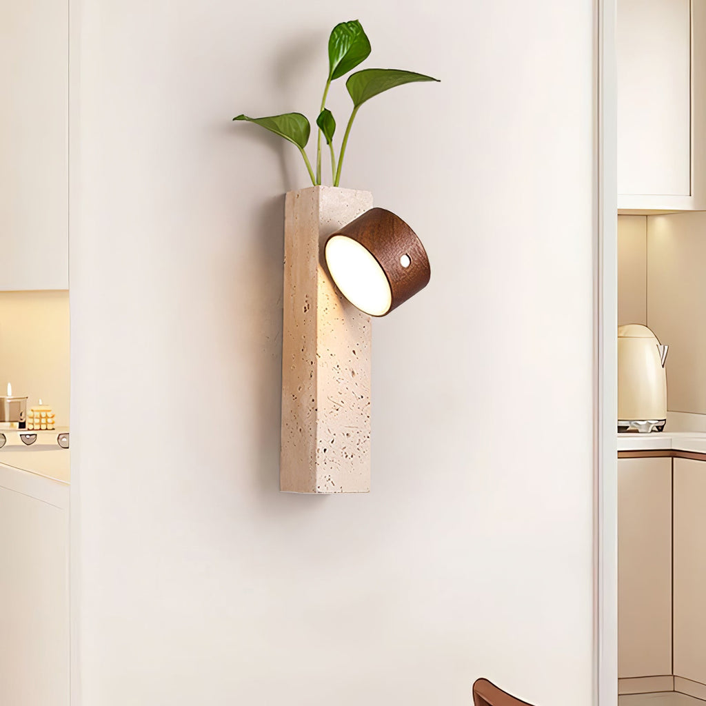 A Clérachic wall light with a dark walnut head tilted on a travertine block, holding a trailing green plant, beside a kitchen doorway.