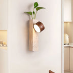 A Clérachic wall light with a dark walnut head tilted on a travertine block, holding a trailing green plant, beside a kitchen doorway.
