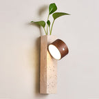 Nearer view of a Clérachic Dark Walnut & Yellow Travertine wall light, its tilted lit head glowing beside a leafy plant.