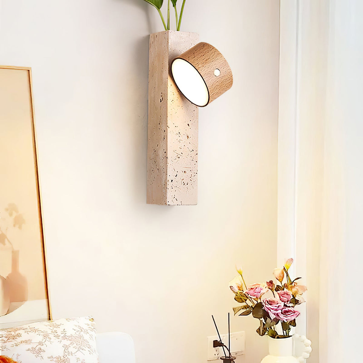 A Clérachic wall light with a tilted walnut head glows on a sunlit wall near a mirror, curtains and a bouquet of pink roses.