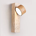 A Clérachic wall light with its pale wood head tilted up and out, showing the lit face above a pitted travertine block.