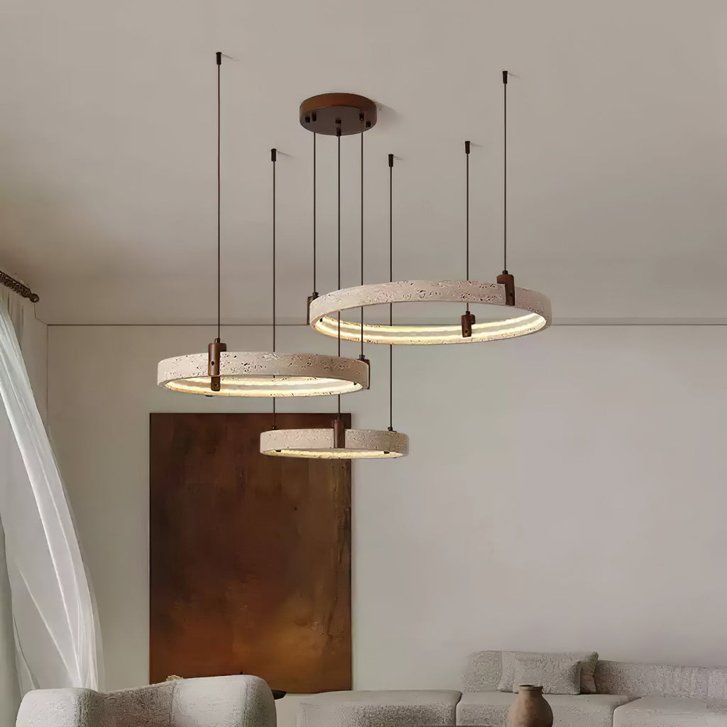 Seen from below, three lit travertine rings from Clérachic hang over a cream sofa, beside a rust-coloured canvas on a pale wall.