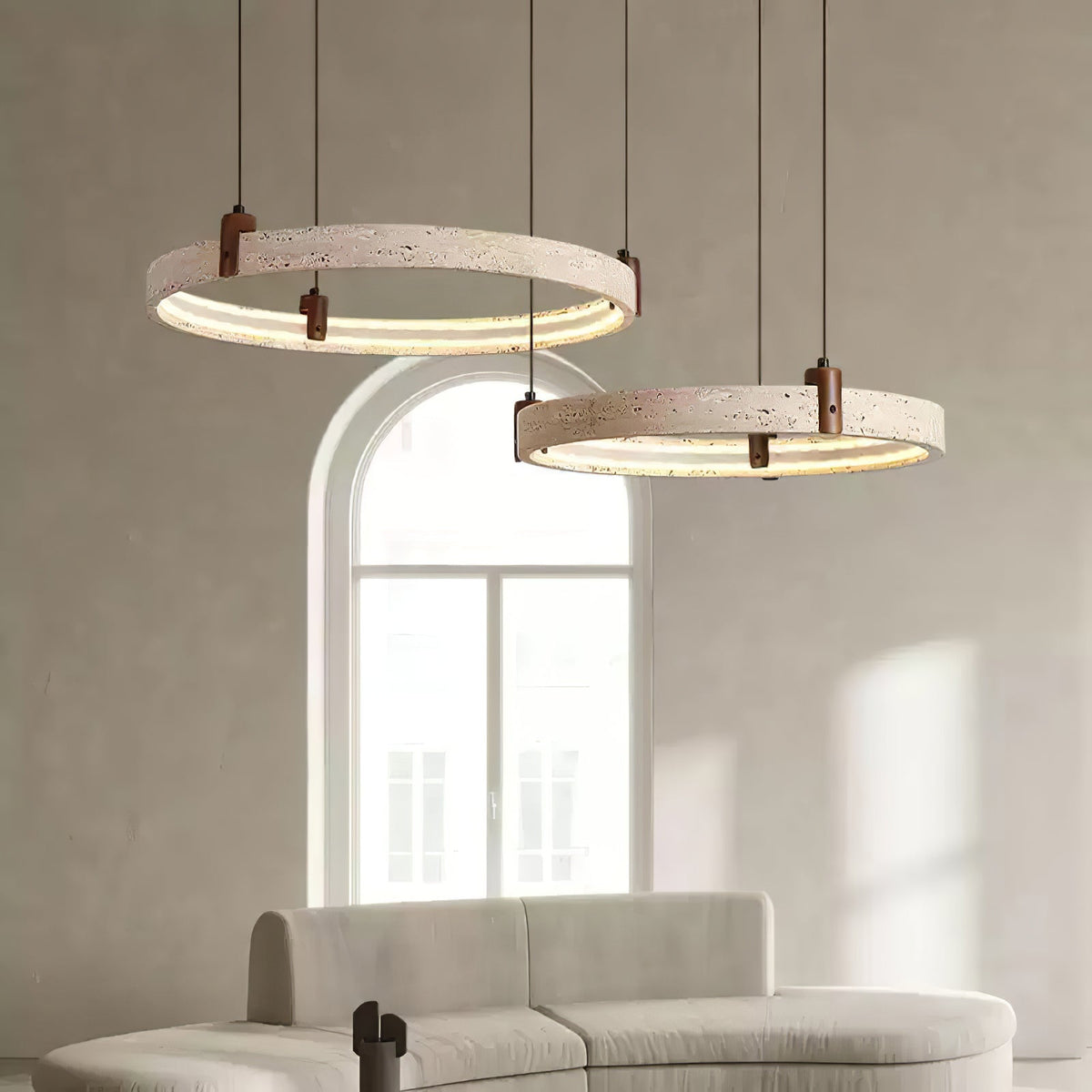 Two of the lit travertine rings on this Clérachic pendant hang in front of an arched window, above the curve of a pale sofa.