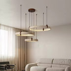 Soft light spills from three travertine rings on a Clérachic pendant beside a sheer-curtained window, above a ruched grey sofa.