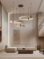 In a tall living room with a stone-toned wall panel and a low timber table, a Clérachic cluster of three travertine rings hangs high.