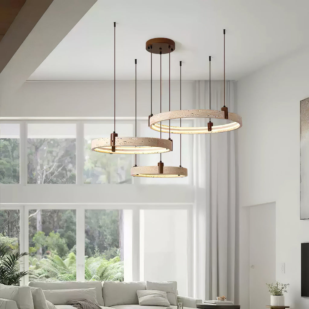This Clérachic pendant of three travertine rings hangs by tall white windows with garden views, above a pale living room sofa.