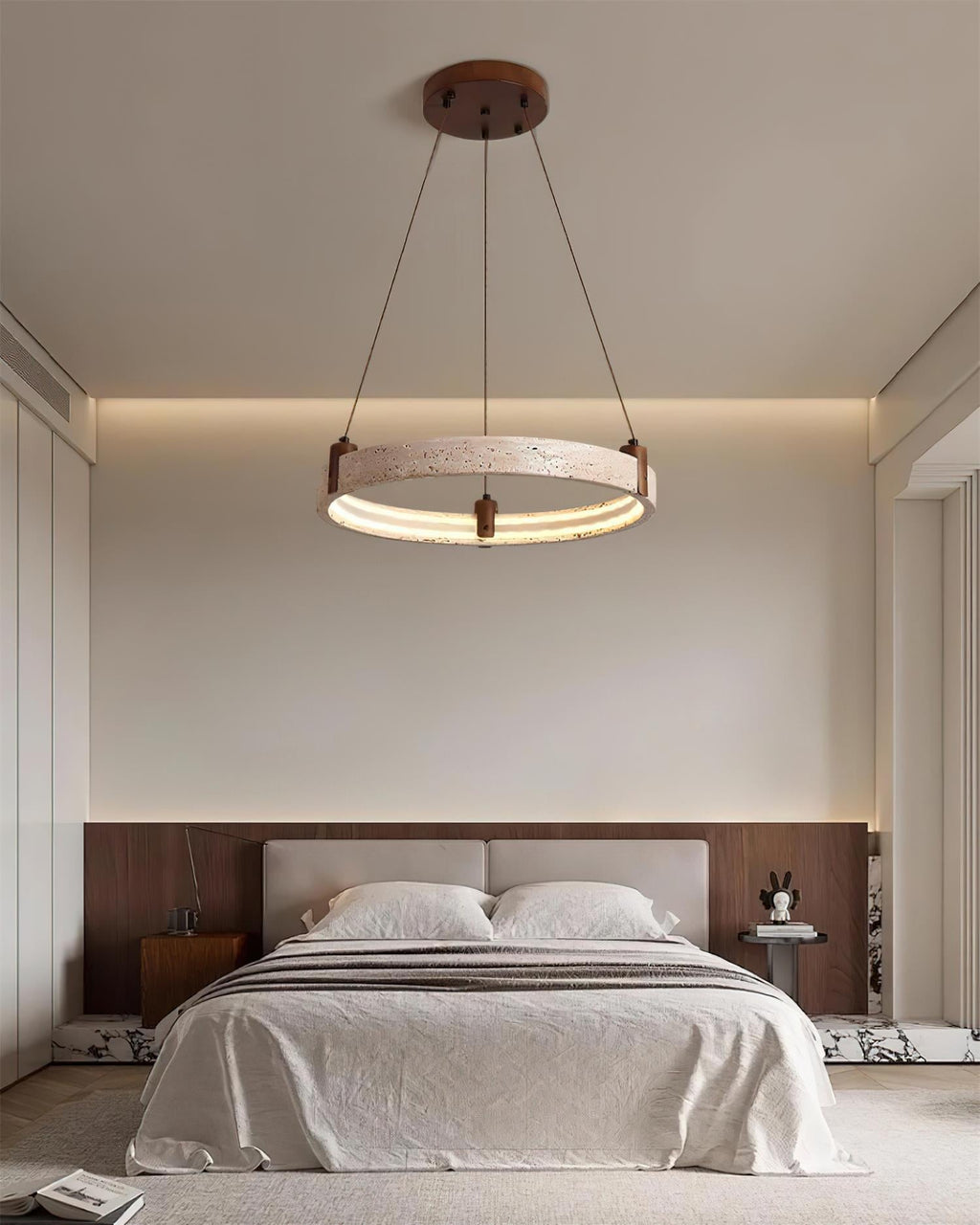 A single Clérachic travertine ring pendant hangs low above a bed in a calm bedroom, with a cove of warm light along the wall.