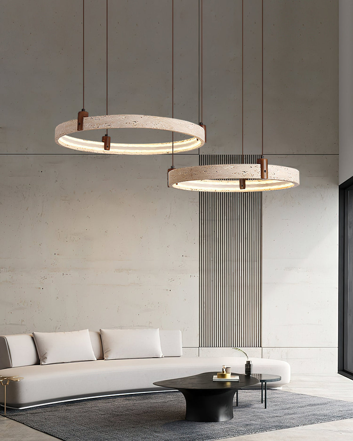 Two travertine rings from Clérachic hang above a low white sofa and black oval table, in front of a concrete wall with slim vertical slats.