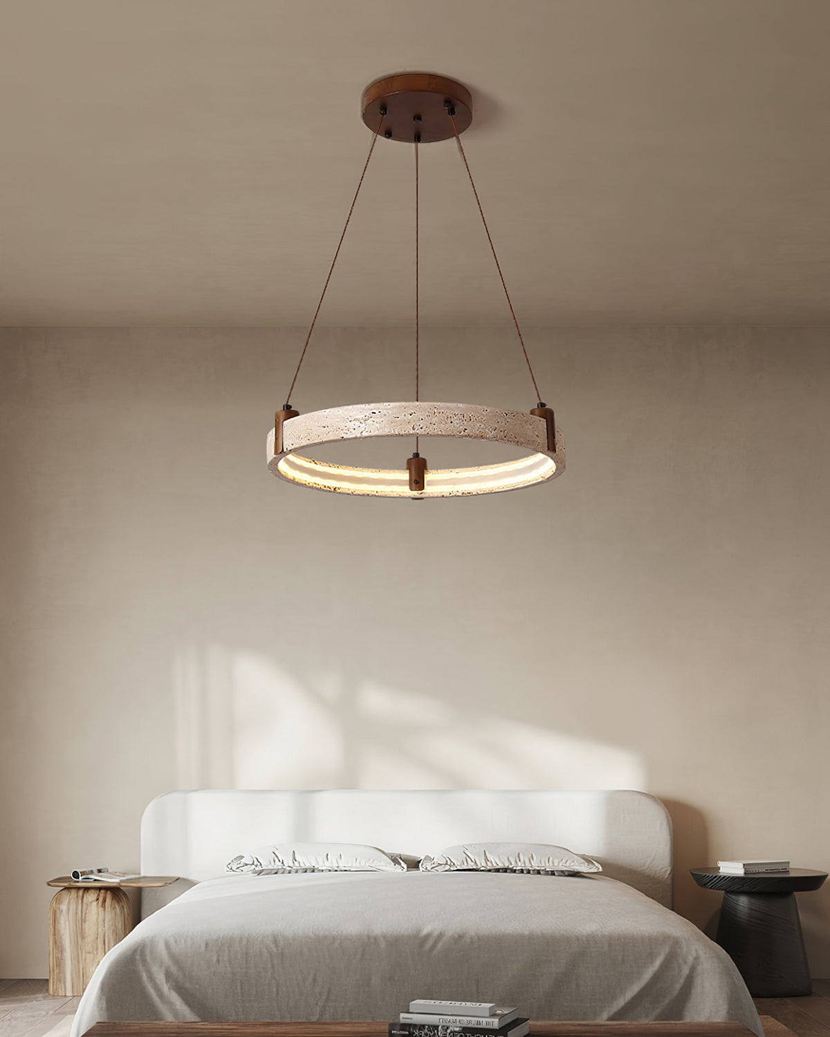 Above a bed with white linen, a Clérachic travertine ring pendant hangs from a round canopy on angled cords, in morning light.