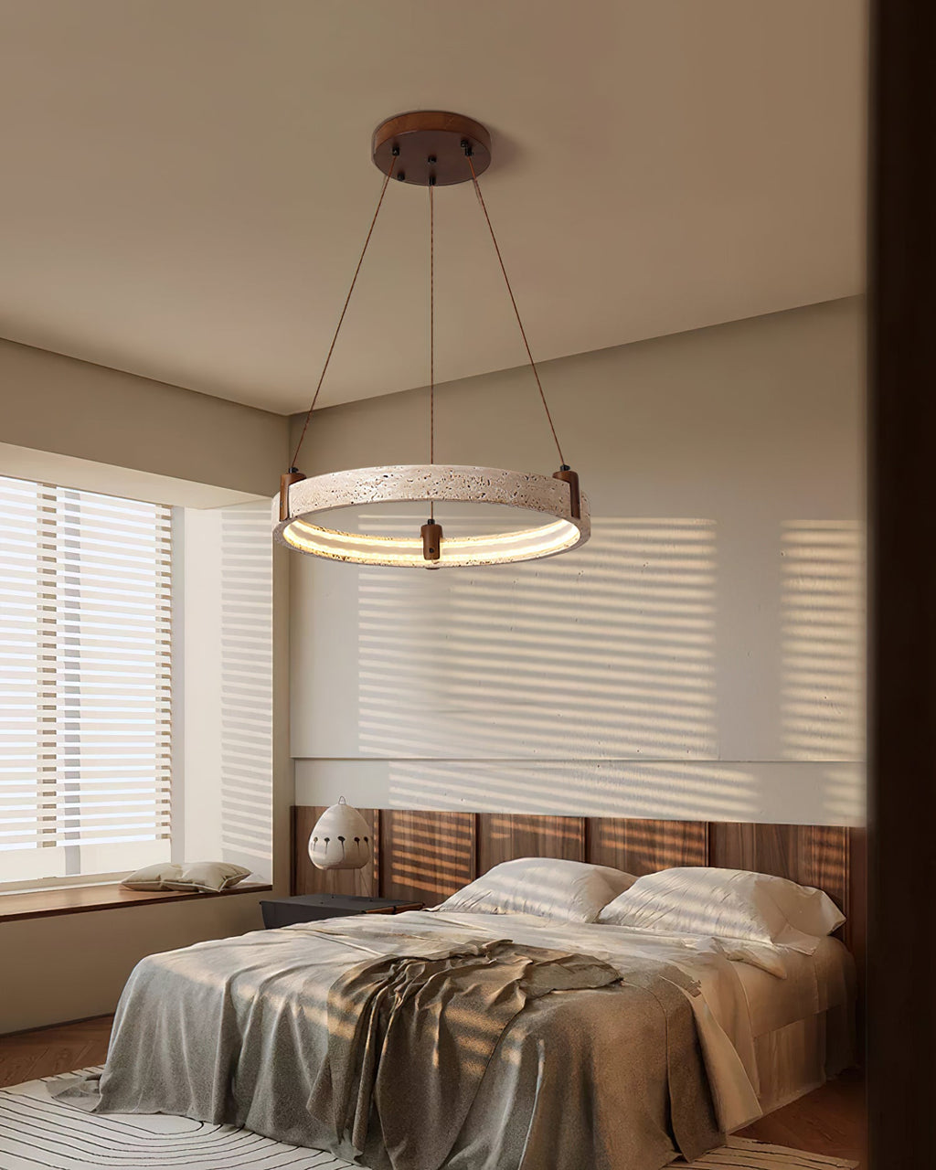 A Clérachic travertine ring pendant hangs above a rumpled bed in a bedroom with slatted blinds and a warm wood headboard wall.