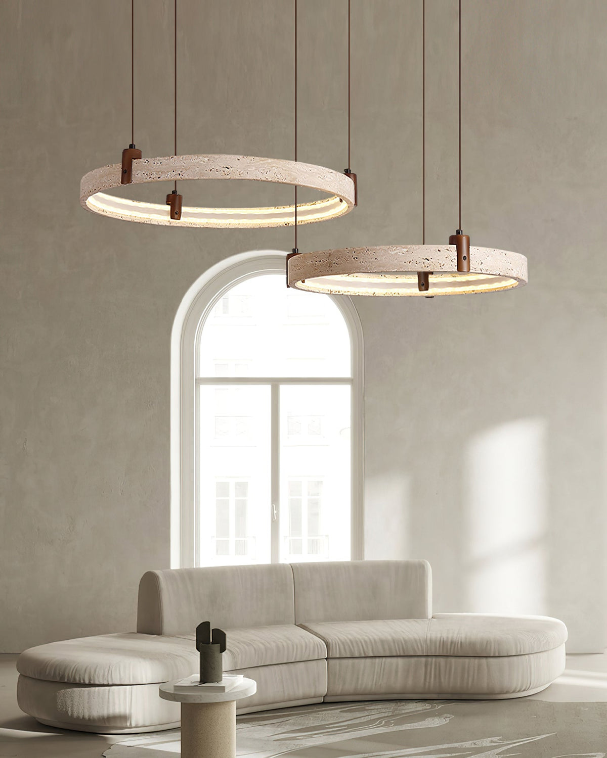 Two travertine rings from Clérachic hang before an arched window, above a pale sofa in a bright, minimal living room.