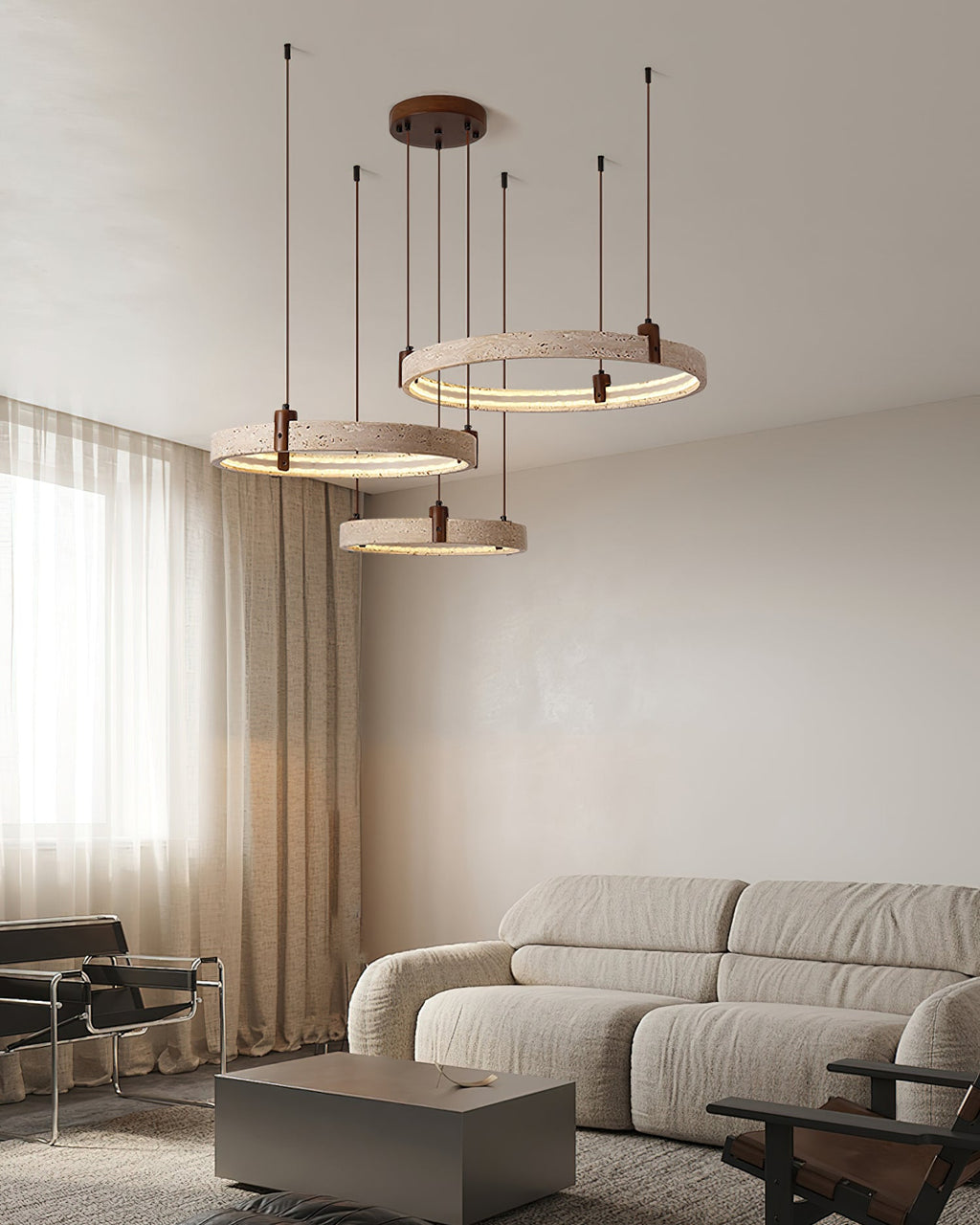 Above a low beige sofa, a Clérachic pendant of three travertine rings hangs by a curtained window, with a grey table below.