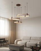 Above a low beige sofa, a Clérachic pendant of three travertine rings hangs by a curtained window, with a grey table below.