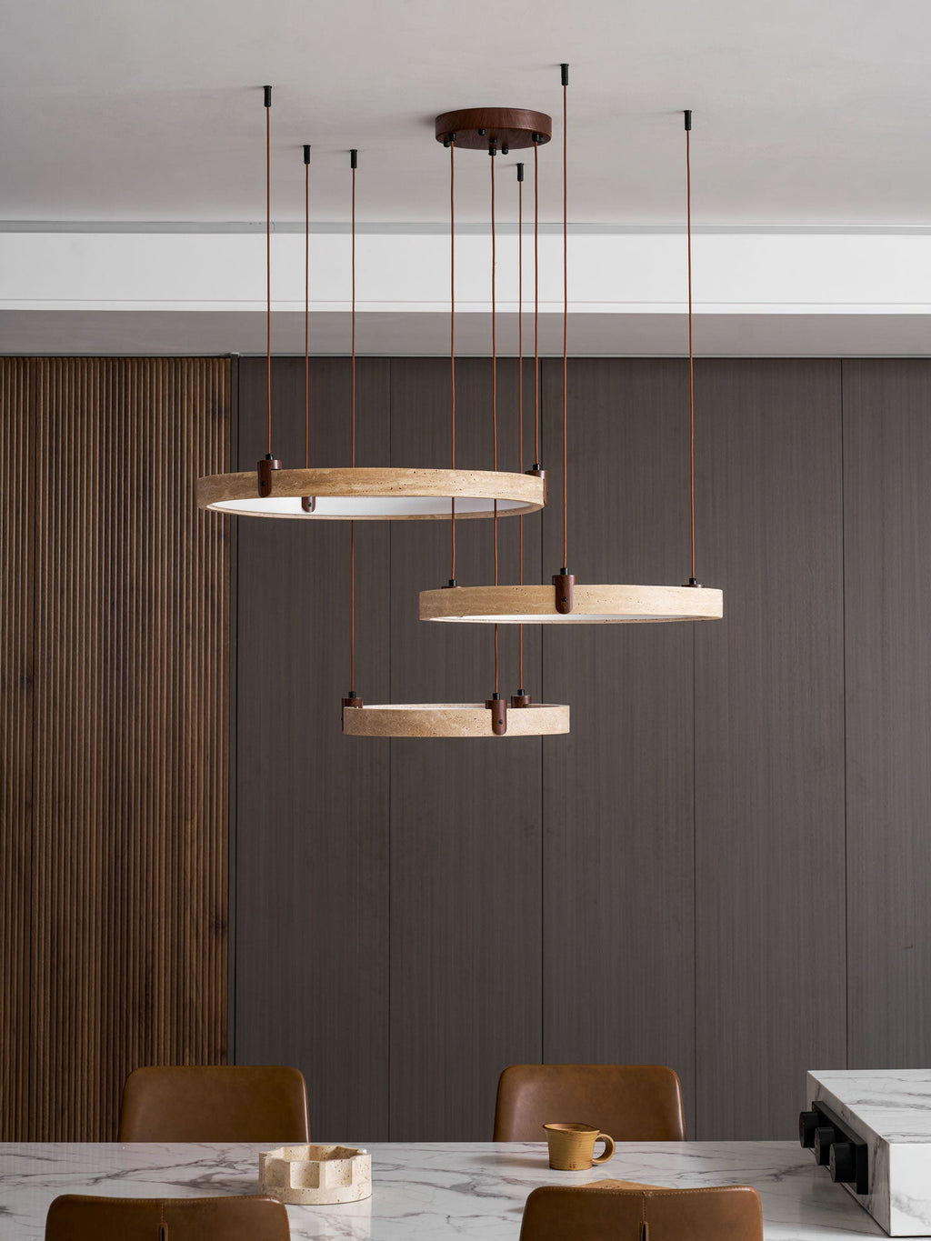 Further back, the three travertine rings of this Clérachic pendant hang over a marble dining table with brown leather chairs.