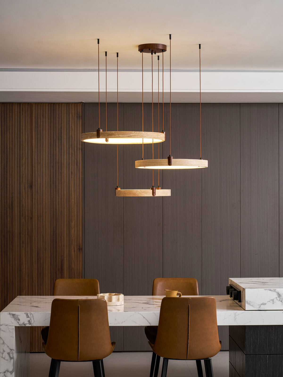 Three golden travertine rings on a Clérachic pendant hang in a modern dining room, with their cords reaching up to a pale ceiling.