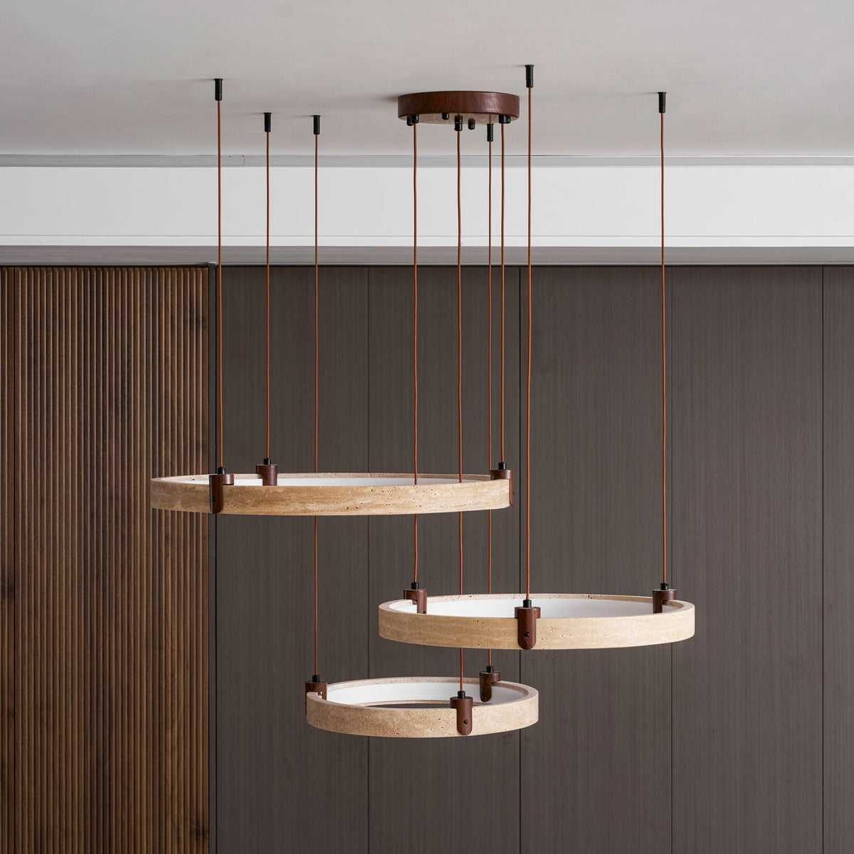 From the side, the three golden travertine rings of this Clérachic pendant hang in front of a dark wood wall.