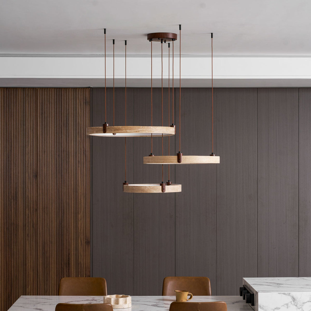 Three golden rings from Clérachic hang before dark wood slats, seen from a distance, over a marble table and tan chairs.