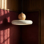 Clérachic speckled travertine disc pendant with a wood ball, hung against deep red fabric panels and warm timber.