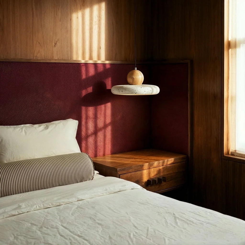 Clérachic travertine disc pendant hung beside a walnut nightstand, with a striped bolster and crisp white bedding.
