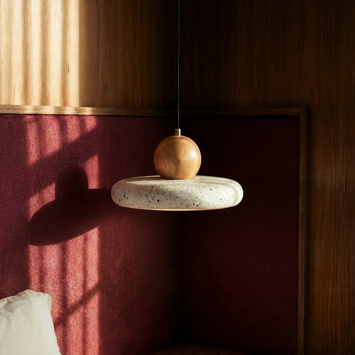 Close view of a Clérachic travertine disc pendant with pitted, speckled stone and a smooth wood ball, against red fabric.