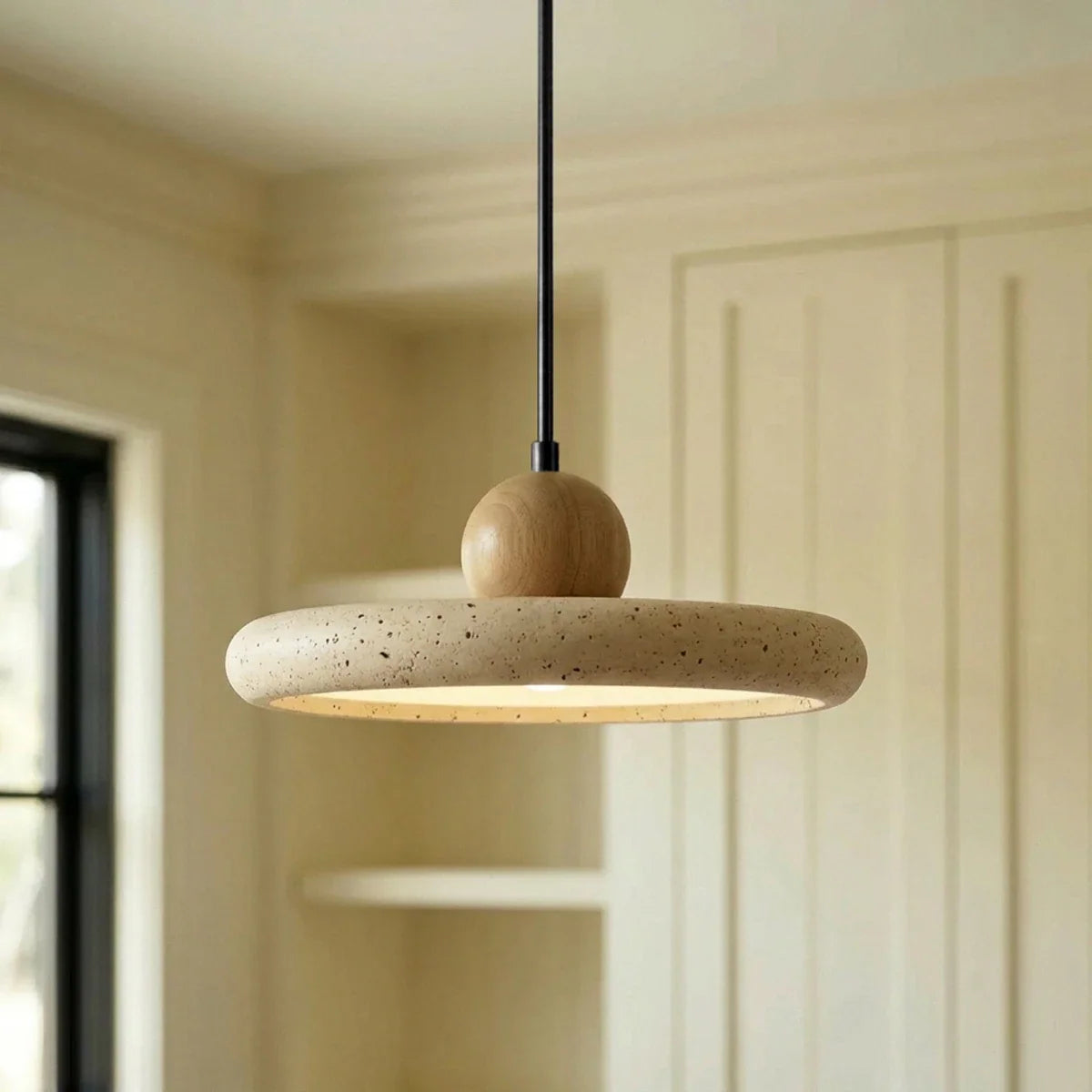 Clérachic travertine disc pendant with a round wood ball on a black stem, lit warm beneath, in a cream panelled room.