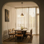 A Clérachic pendant light hangs over a wooden dining table with cane-back chairs in a sunlit arched alcove.