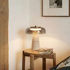 Lit Clérachic table lamp with a smoky grey dome on a wooden stool beside a framed print, with a few pebbles and a book nearby.