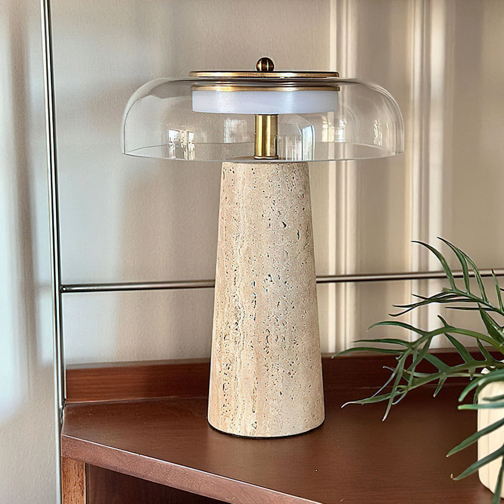 A clear glass dome over a tall travertine base, this Clérachic lamp sits on a dark wood sideboard with a green plant to the right.